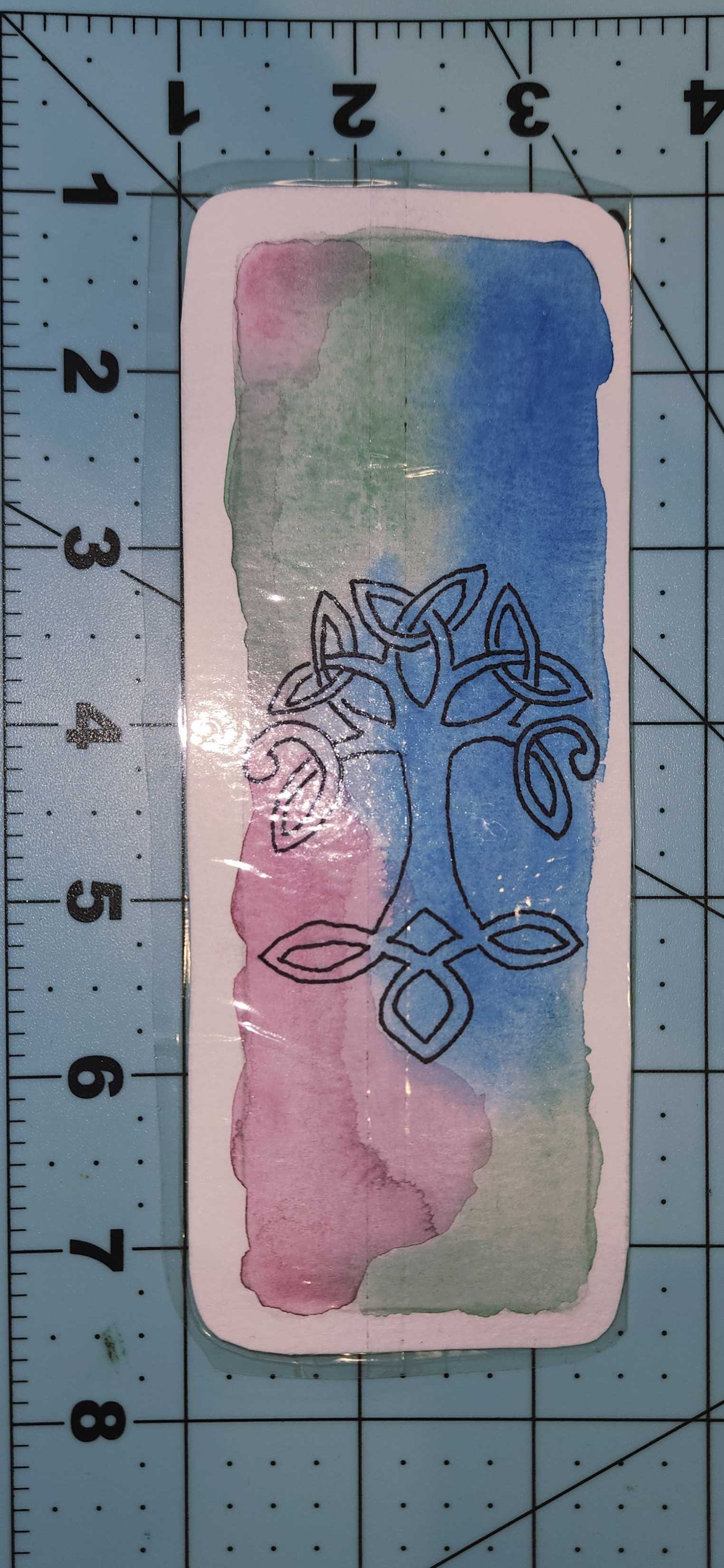 Decorative card with Celtic knot design on a watercolor background, placed on a cutting mat with measurements.